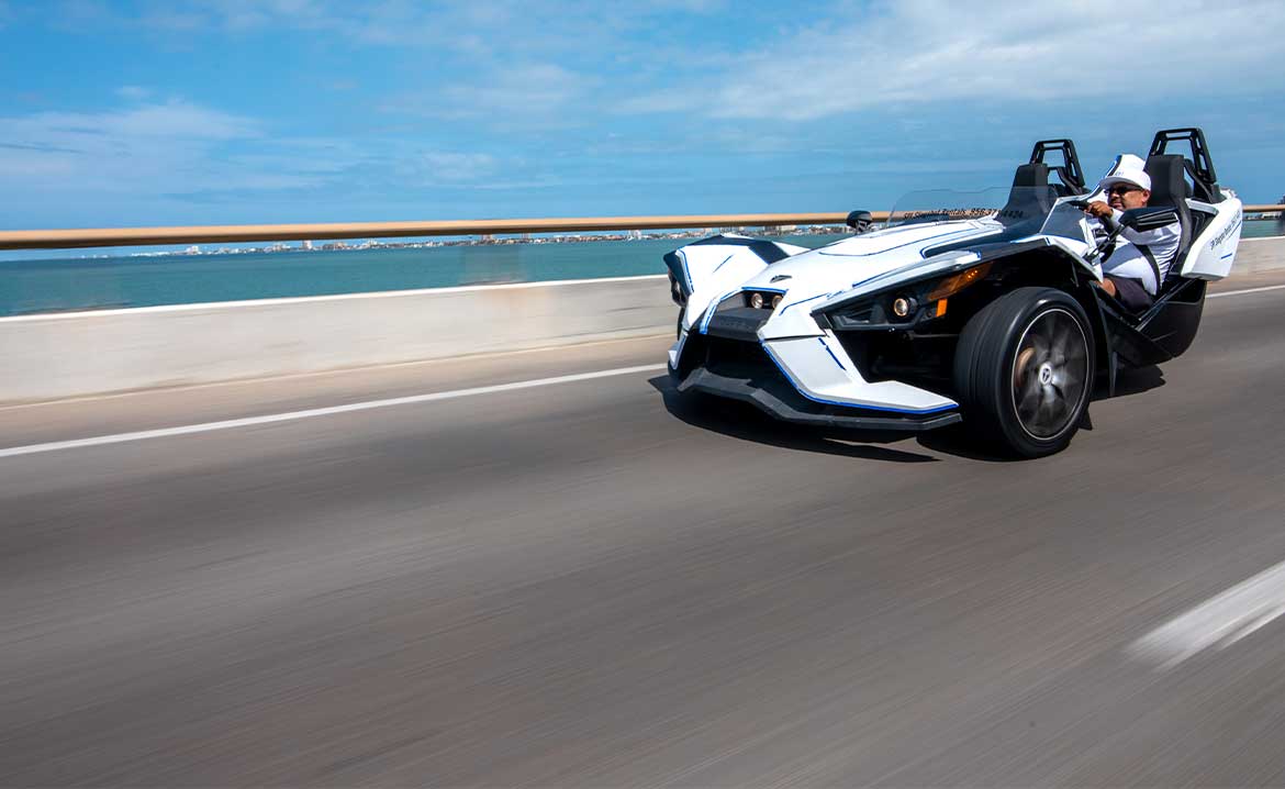 A man driving a slingshot Spyder along a sunny highway in South Padre Island.