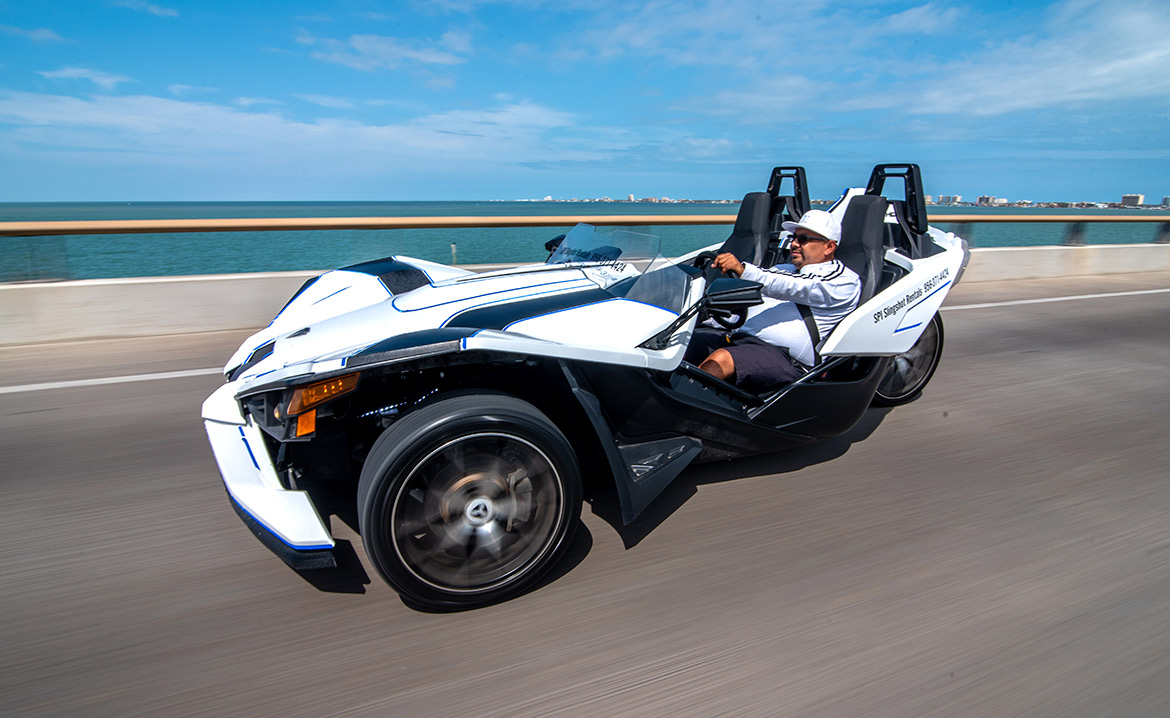A man on a black and white slingshot speeds along the highway, heading towards South Padre Island for an adventure.