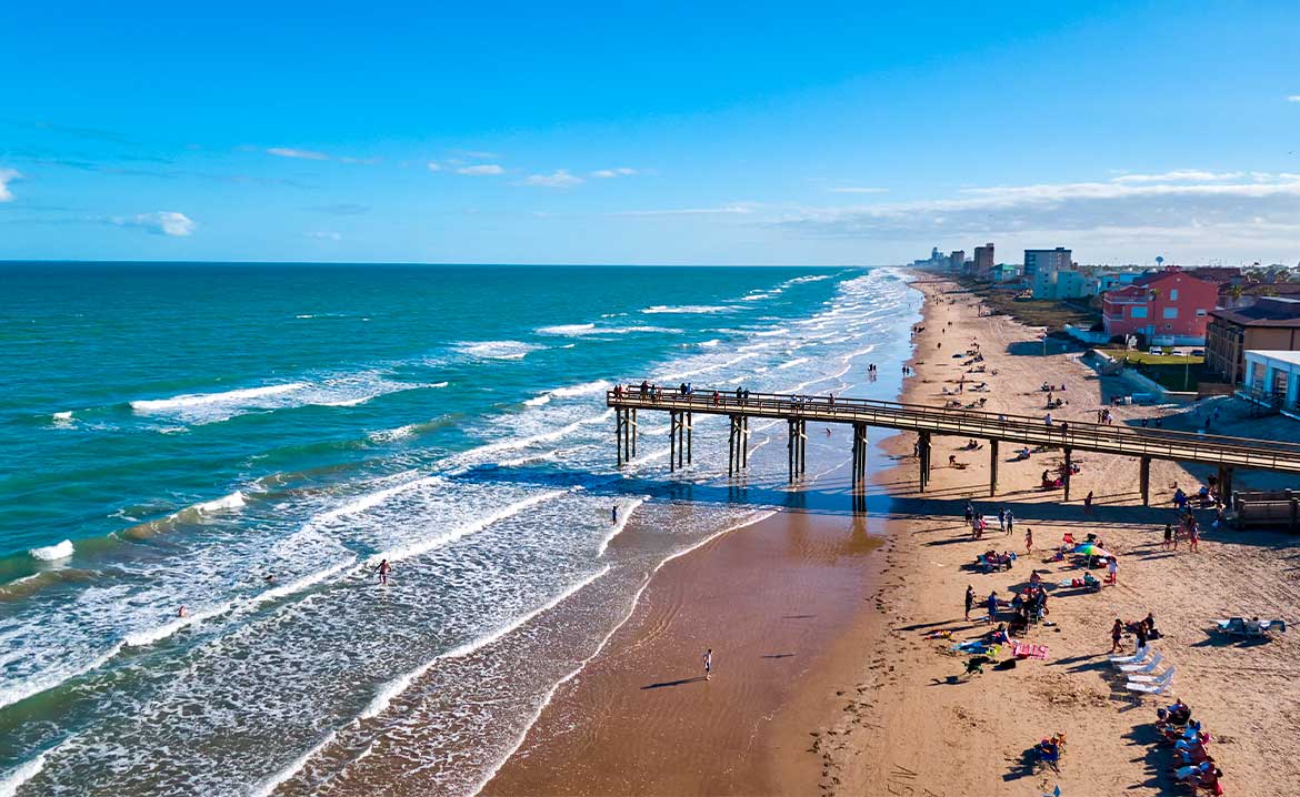 South Padre Island beach scene featuring people on the sand and a wooden pier in the background under a clear sky.