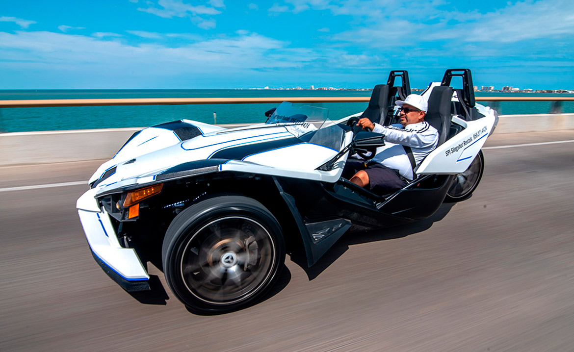 A man on a black and white motorcycle cruising down the road, advertising slingshot rentals in South Padre Island.