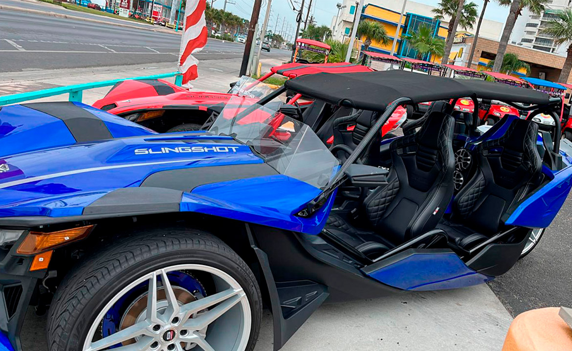 A blue and black sports car parked along the road, advertising slingshot rentals in South Padre Island.