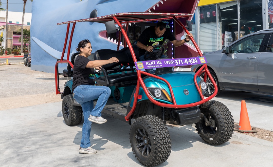 A woman pushes a golf cart featuring a shark design on the side, set against a green golf course background.